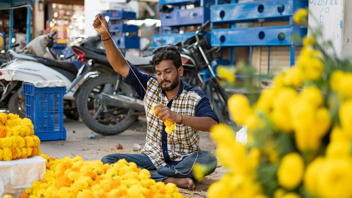 A man sitting on a busy street stringing yellow marigold flowers into a garland