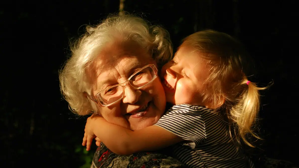 A smiling grandmother with white hair and glasses hugging a young grandchild