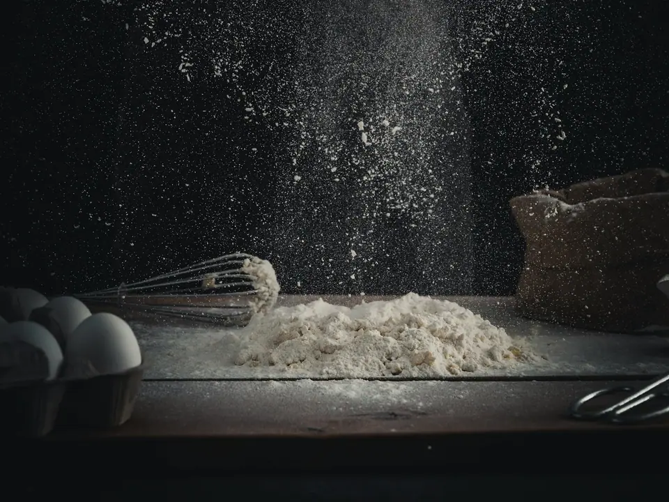Flour being poured onto a table beside eggs and a whisk