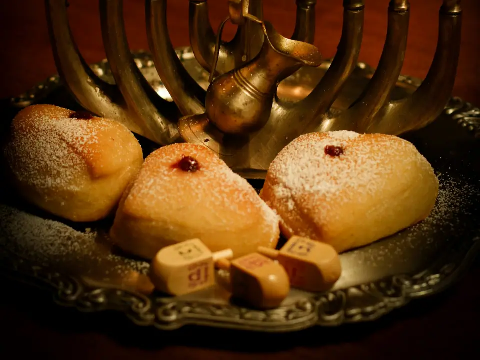 Wooden dreidels and doughnuts on a tray beside a brass menorah