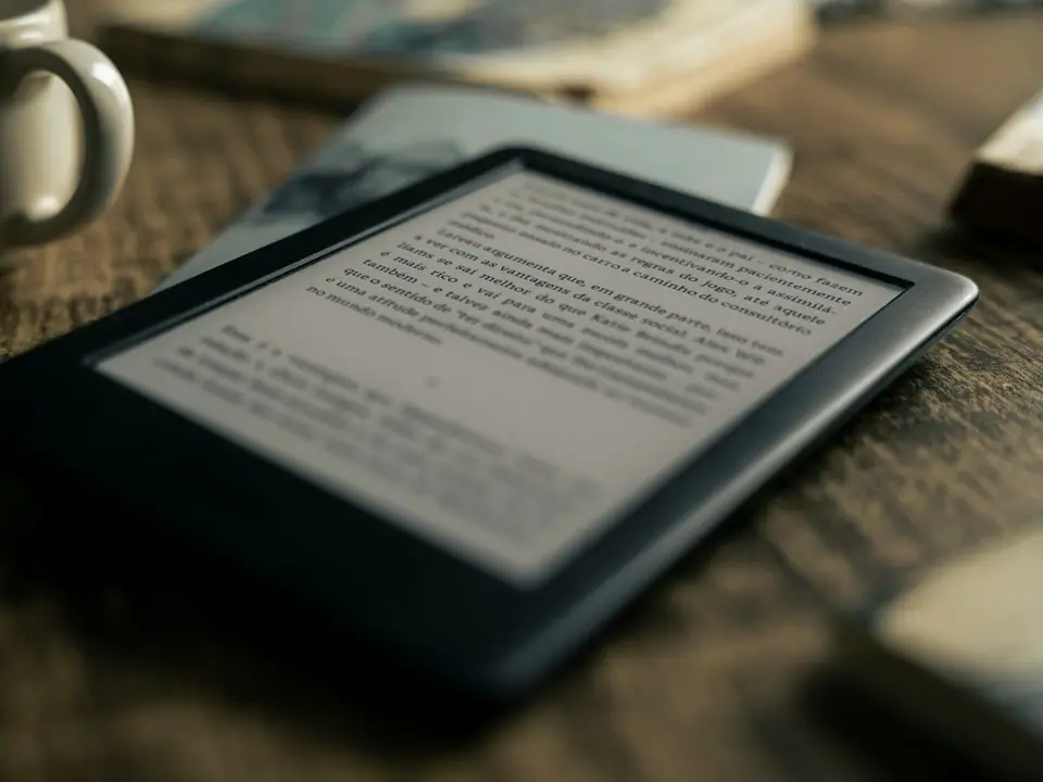 An e-reader on a wooden table beside coffee and books