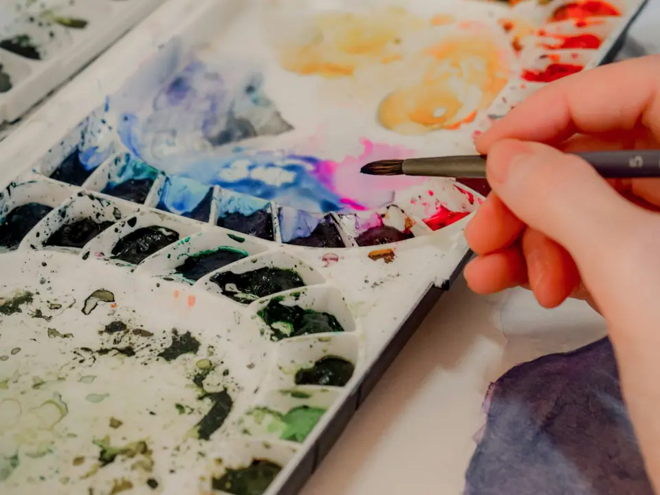 A hand dipping a brush into watercolor paints during a painting lesson