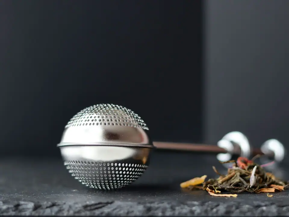 A round stainless steel tea infuser ball beside loose tea leaves
